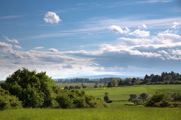 Blick auf eine Mittelgebirgslandschaft mit Wiesen, Feldern und Bäumen. Am Horizont ist eine Bergsilhouette zu sehen mit einem Sendeturm. Das Wetter ist sonnig und schön.