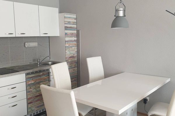Ein weißer Esstisch mit fünf Stühlen steht vor einer weißen Küchenzeile. Die Kühl- und Hochschranktüren sind bunt gestreift. Eine runde, graue Lampe hängt über dem Tisch.
