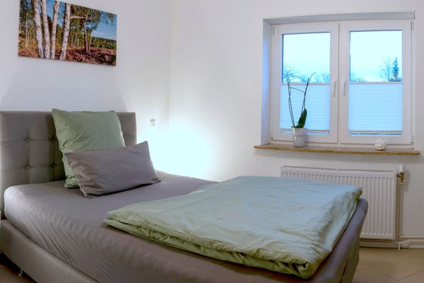 Das Bild zeigt ein gemütliches Schlafzimmer. Ein graues Polsterbett mit passender grauer Bettwäsche und grünen Kissen dominiert den Raum. Über dem Bett hängt ein Bild, das eine Waldlandschaft mit Birkenbäumen zeigt. Rechts ist ein Fenster mit weißen Jalousien, auf dessen Fensterbank eine Pflanze und Dekoration stehen. Helle Fliesen bedecken den Boden, und die Wände sind weiß gestrichen.
