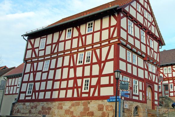 Außenansicht eines viergeschossigen Fachwerkhauses mit rot-braunen Balken und großer, abgerundeter Eingangstür aus hellem Holz auf der Stirnseite. Der Sockel ist aus Sanstein, ein Kellerabgang führt zu einer großen Tür mit halbrundem Glaseinsatz als oberem Teil.