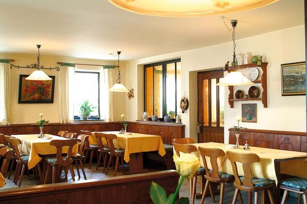 Ein Gastzimmer im ländlichen Stil mit Holzbankrückwänden, die mit Schnitzereien verziert sind. Zwischen Bänken und Stühlen stehen gedeckte Tische. Bilder und ein Schüsselkorb zieren die Wände. Vor den Fenstern hängen helle, gemusterte Vorhänge mit grüner Borte.