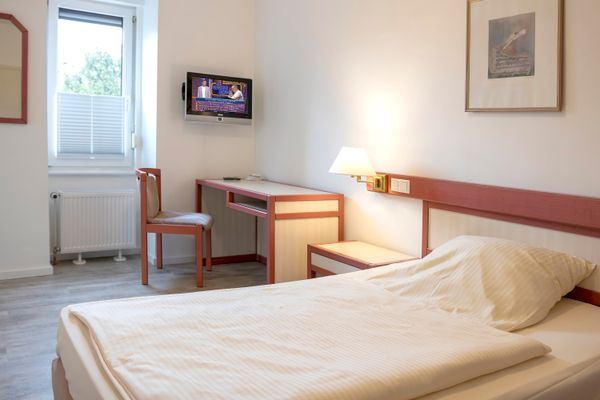 Das Bild zeigt ein Hotelzimmer mit einem Einzelbett, das mit weißer Bettwäsche bezogen ist. Neben dem Bett befindet sich ein Nachttisch mit einer Wandlampe darüber. Links steht ein Schreibtisch mit einem Stuhl, darüber hängt ein kleiner Fernseher an der Wand. Ein Fenster mit halb heruntergelassenem Rollo und ein Heizkörper sind ebenfalls zu sehen. Der Boden ist mit hellem Laminat ausgestattet.