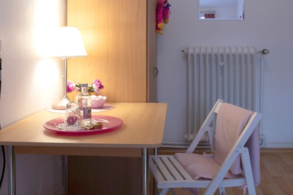 Kleiner Tisch mit Lampe, Glas und Flasche, daneben ein Stuhl und ein Heizkörper im Hintergrund