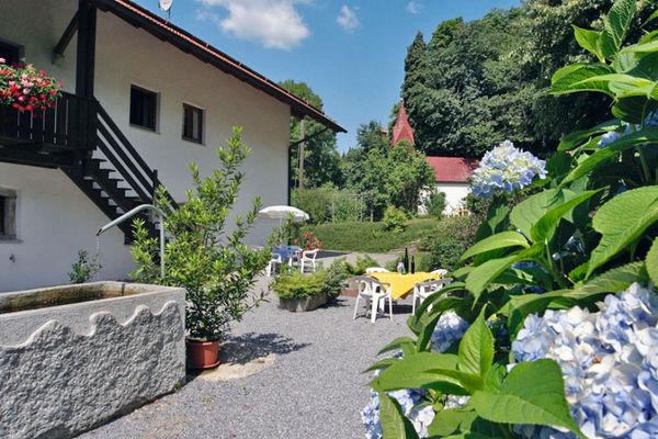 Auf einer kiesbestreuten Terrasse stehen zwei Tischgruppen mit je vier Stühlen vor einem Haus mit Treppenaufgang zum Balkon. Im Vordergrund ein großer Steintrogbrunnen und eine blau blühende Hortensie. Im Hintergrund steht verdeckt hinter Bäumen und Sträuchern eine Kapelle.