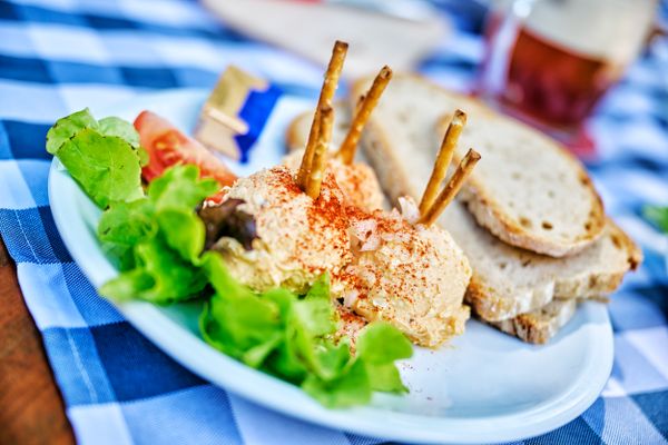Auf einer Blau-weiß karierten Tischdecke steht ein Teller mit einer Portion Obatzda mit ein paar Scheiben Brot. Das Ganze ist garniert mit Tomate, Salat, und Butter. Im Hintergrund steht ein dunkles Bier.