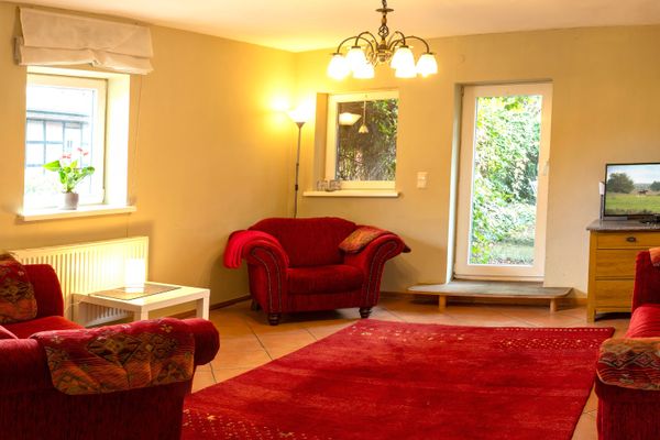 Ein Wohnzimmer mit gelblich gestrichenen Wänden, Terrakotta-Fliesen und einem großen roten Teppich in der Mitte. Im Raum stehen rote Polstersessel, ein weißer Couchtisch und ein Holzschrank mit einem Flachbildfernseher. Mehrere eingeschaltete Lampen sowie Tageslicht aus zwei Fenstern und einer Glastür erhellen den Raum.