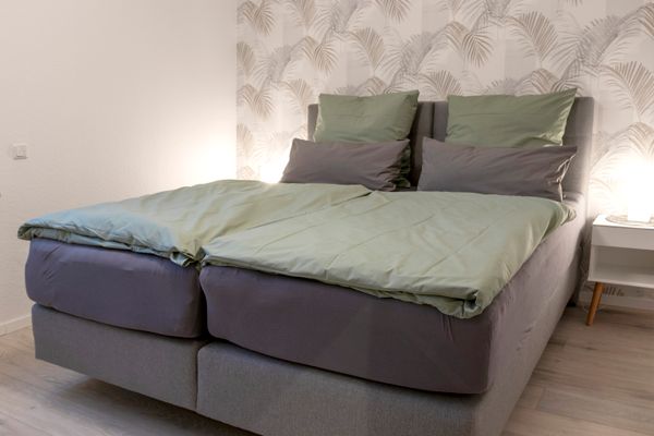 Das Bild zeigt ein Schlafzimmer mit einem Doppelbett. Es besteht aus zwei separaten Matratzen, bezogen mit grauen Spannbettlaken. Die Bettdecken sind hellgrün, und auf jeder Seite liegen zwei Kissen: ein großes hellgrünes und ein kleineres graues. Im Hintergrund schmückt eine Tapete mit dezentem Palmenmuster die Wand. Rechts steht ein Nachttisch mit einer eingeschalteten Lampe, die warmes Licht verbreitet. Der Boden ist mit hellem Holzlaminat ausgelegt.