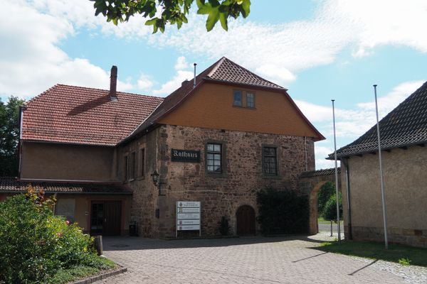 Ausschnitt eines braunen Fachwerkhauses mit Sandsteinsockel. eine einfache, mit Efeu berankte Treppe führt zur schlichten Eingangstür, die oberhalb ein geschnitzter Segensspruch ziert. Es steht geschrieben: Lebe und Liebe, für dieses Haus.