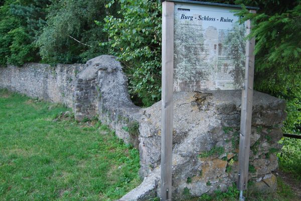 Eine Bruchsteinmauer umschließt eine Wiese, dahinter stehen hohe Bäume. Vor der Mauer befindet sich ein Schild, das Informationen über die Burg-Schloss-Ruine erteilt.