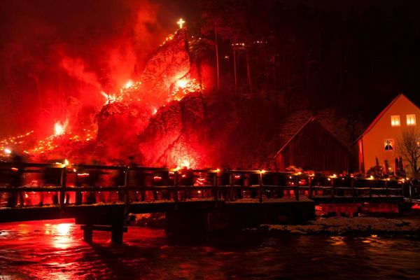 Besucher des Lichterfestes laufen über eine Holzbrücke bei Nacht. Im Hintergrund wir dein Felsen durch bengalische Fackeln zum Leuchten gebracht.