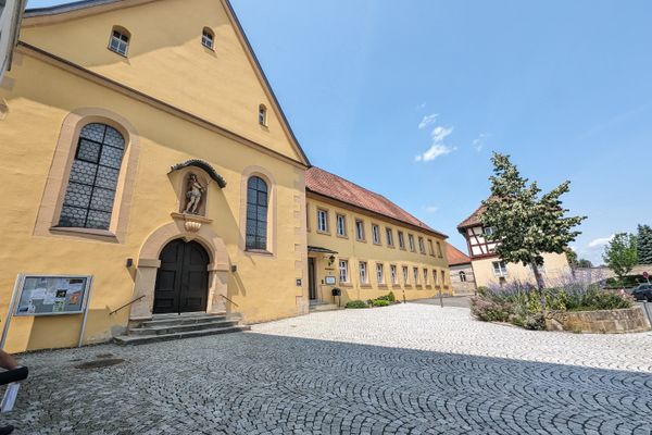 Das Bild zeigt die Außenansicht des Oblatenklosters St. Heinrich in Kronach. Das Gebäude trägt ein hellbraune Farbe. Rechts daneben sieht man einen kleinen Garten.