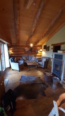 Hoher Wohnraum in einem Holzhaus mit grauer Sitzecke, einem Tisch und einem Kaminofen mit Sichtscheibe.