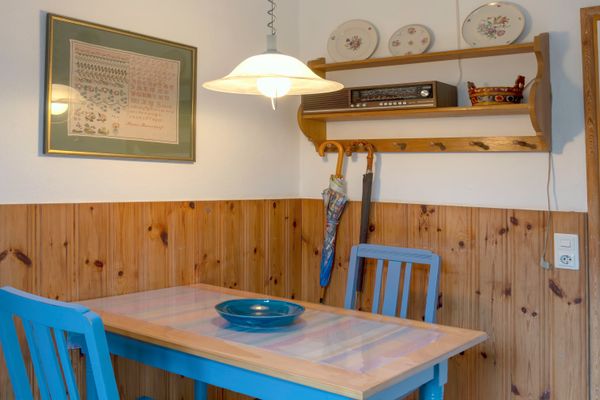 Kleiner Tisch mit zwei Stühlen vor einer holzverkleideten Wand, darüber Regal und Hängelampe.