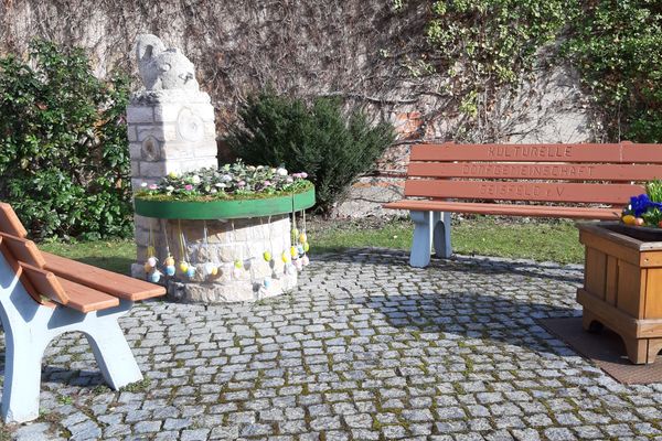 Ein kleiner, gepflasterter Platz mit zwei Ruhebänken, zwischen denen ein kleiner Brunnen aus hellem Gestein steht. Auf dem Wasserbecken sind Nadelzweige mit bunten Eiern zu sehen.