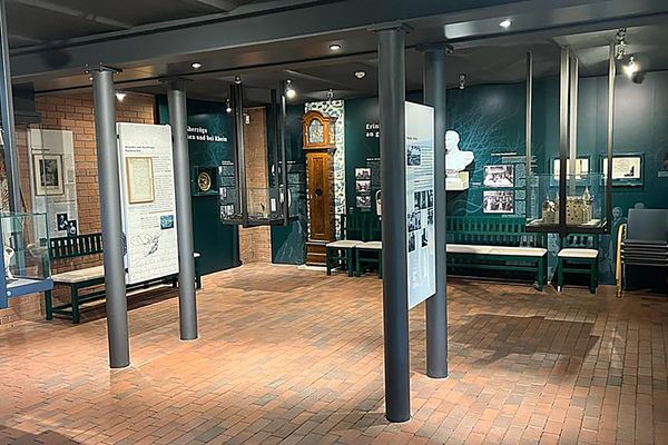 Blick in einen Ausstellungsraum mit Stahlträgern an der Decke und markanten Stahlsäulen, die diese tragen. Dazwischen hängen Infotafeln, genauso an den Wänden. Rings um sind Exponate ausgestellt.