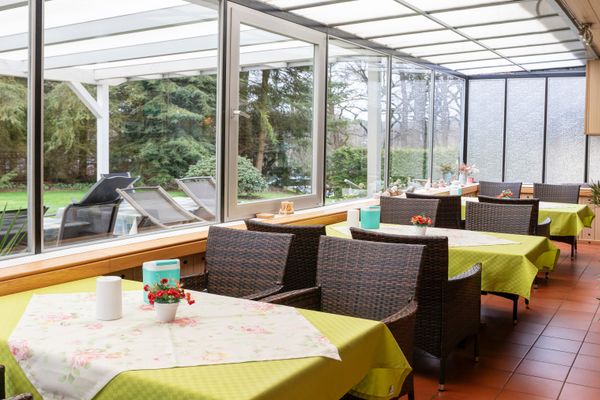 Wintergarten mit Tischen und Stühlen, großen Fenstern und Blick in den Garten, Tageslicht.