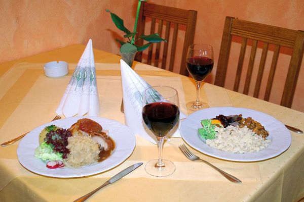 Auf dem gedeckten Tisch stehen zwei Gerichte auf weißen Tellern, daneben Servietten, zwei Gläser Rotwein und eine kleine Blumenvase mit einer weißen Rose.