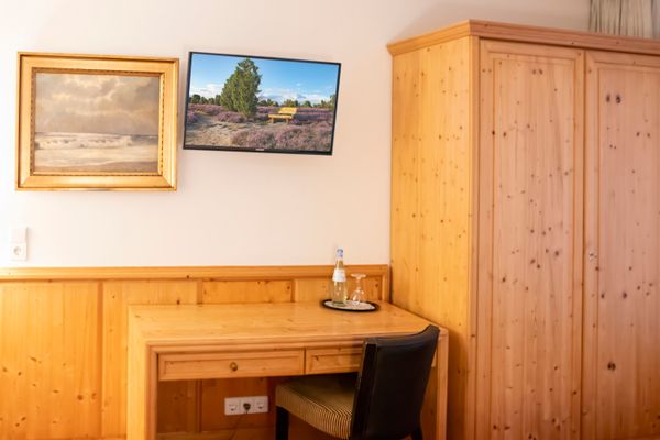Das Bild zeigt einen Raum mit Holzmöbeln, einem Gemälde mit Meereslandschaft und einem Flachbildfernseher, der eine Landschaft mit Bäumen und Blumen zeigt. Ein Schreibtisch mit Wasserflasche und Glas steht unter dem Fernseher, rechts ist ein großer Holzschrank.