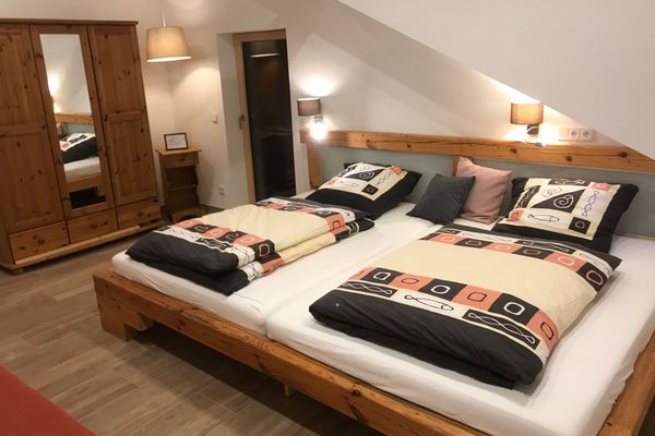 Unter einer hellen Mansarde steht ein breites Doppelbett mit Holzrahmen und Nachttischlampen mit rundem Schirm. Daneben ein Schrank mit Spiegeltür und ein zusätzliches kleines Holzregal mit Schublade.