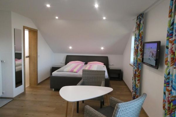 Auf einem Holzboden steht ein graues Boxspringbett mit Nachkonsolen unter einer hellen Dachschräge. Strahler leuchten von der Decke. An der Wand, zwischen bunten Vorhängen, hängt ein Flachbildfernseher. Davor stehen ein Tisch und zwei Sessel.