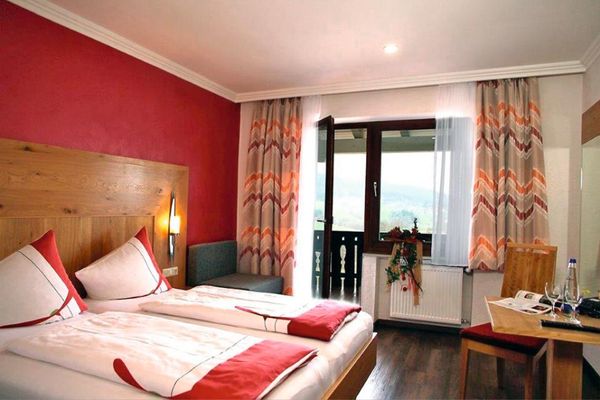 Ein Doppelbett mit halbhoher Holzrückwand und einer rot gestrichenen Wand hinter dem Bett. Gegenüber befinden sich ein Tisch, ein Stuhl und ein Spiegel. Die Tür zum Balkon steht offen, helle, rot-orange gemusterte Vorhänge rahmen Fenster und Balkontür.