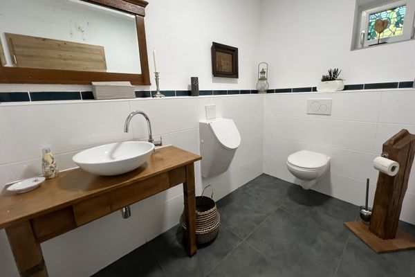 Badezimmer mit Waschtisch, großem Spiegel und Toilette. Die Fließen sind grau und weiß. Das Möbel ist aus Holz.