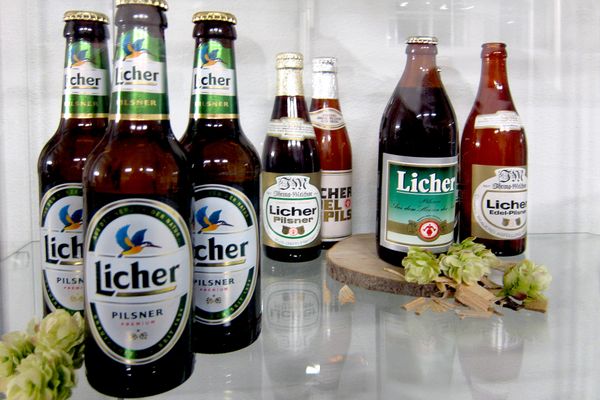 Blick in eine Glasvitrine, in der mehrere Bierflaschen mit Etiketten der Licher Brauerei ausgestellt sind. Manche Etiketten tragen im Logo einen Eisvogel, andere ein sogenanntes rotes Hessenmächen. Zur Dekoration liegen ringsum Hopfenblüten.