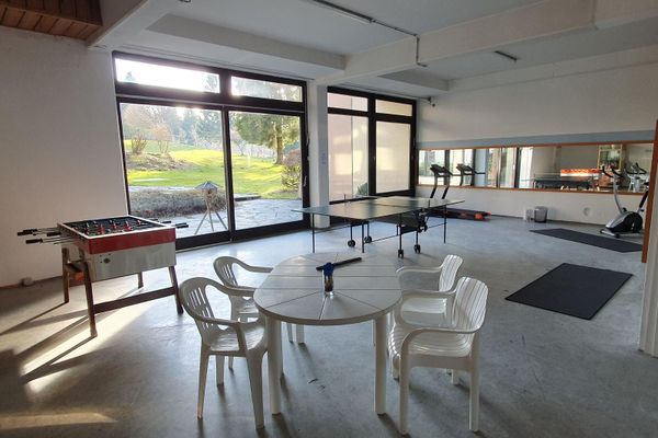 Vor der großen Fensterfront stehen ein Kickerkasten, eine Tischtennisplatte, ein weißer, ovaler Gartentisch mit vier Stühlen, ein Laufband und ein Ergometer. Im Hintergrund ist ein weiterer Fitnessraum erkennbar.