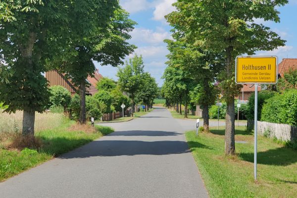 Gerade Dorfstraße mit beidseitigen Bäumen, Wohnhäusern und Gärten, rechts gelbes Ortsschild „Holthusen II – Gemeinde Gerdau – Landkreis Uelzen“, Blick entlang der Fahrbahn unter blauem Himmel mit vereinzelten Wolken.