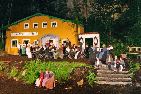 Vor einem gelben Gebäude mit grünem Dach, über dem „Zum Leitner Wirt“ steht, herrscht lebhaftes Treiben. Auf der Terrasse und dem Gelände davor sitzen und stehen zahlreiche Gäste, auch Kinder sind unter ihnen.