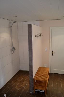 Begehbare, offene, gemauerte Dusche mit hellen Wand- und dunklen Bodenfliesen; an der Duschmauer vier Garderobenhaken, davor eine Holz-Badezimmerbank mit Ablage.