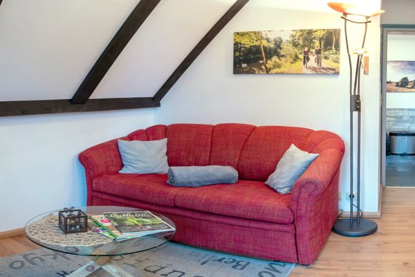 Rotes Sofa mit Kissen vor schräger Wand mit sichtbaren Balken, davor ein runder Glastisch