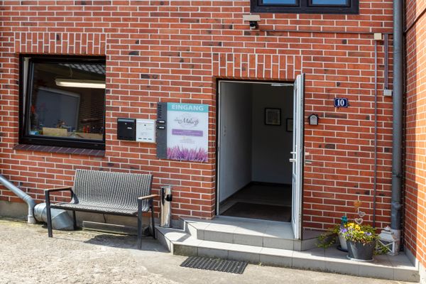 Eingang eines Backsteingebäudes mit geöffneter Tür, Sitzbank, Blumentöpfen und Hinweisschild neben der Tür.