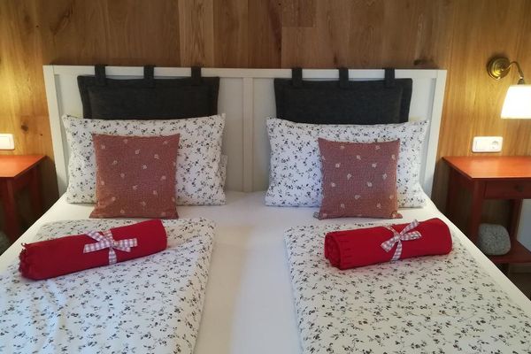 Vor einer Holzwand steht ein Doppelbett mit weißer Rückwand, zwischen zwei roten Nachttischen. Diese werden von Wandlampen beleuchtet. Auf dem Bett liegen, umwickelt von roten karierten Bändern, zwei rote, gerollte Wolldecken.