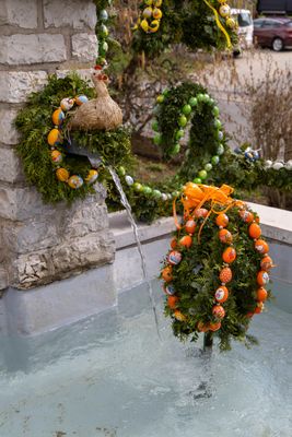Strohhuhn auf Wasserspeier am Brunnen. Um den Wasserspeier Kranz mit bemalten Ostereiern. Im Brunnentrog Girlanden in Eiform mit Ketten aus bunt bemalten Eiern geschmückt.