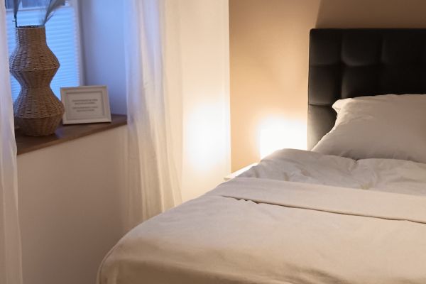 Bett mit weißer Bettwäsche, neben dem Bett ist eine Lampe, die das Schlafzimmer in ein gemütliches Licht taucht. Auf der Fensterbank neben dem Bett steht eine Vase mit Trockenblumen und ein kleiner Bilderrahmen.