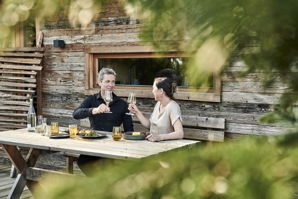 Ein Paar mittleren Alters sitzt an einem Holztisch vor einer Holzhütte im Freien, umgeben von Grün. Sie prosten sich mit Weingläsern zu und auf dem Tisch steht Essen vor ihnen.