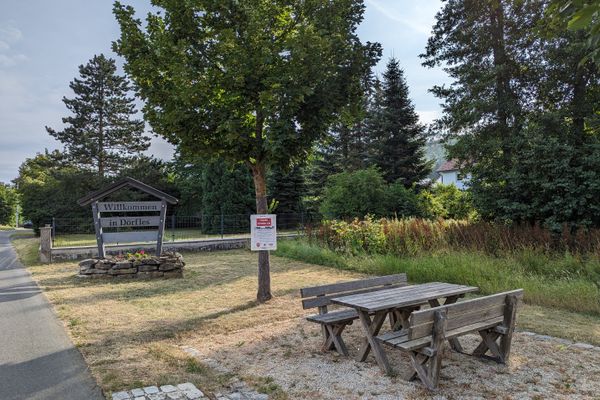 Das Bild zeigt einen Rastplatz am Ortseingang in Dörfles. Man sieht eine Sitzgruppe bestehens aus 2 Bänken und einem Tisch in der Mitte. Dahinter erkennt man das Ortsschild des Ortes Dörfles. Des Weiteren sieht man Bäume im Hintergrund.