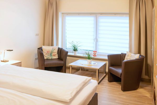 Sitzecke in einem hellen Zimmer mit zwei braunen Sesseln, kleinem Holztisch, Zimmerpflanzen auf der Fensterbank und beige Vorhängen.