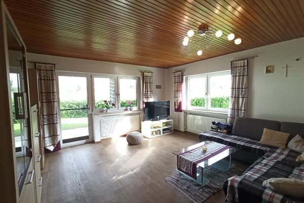 In einem großen Wohnzimmer mit Holzdecke steht ein graues Ecksofa mit einer karierten Decke auf den Sitzflächen, ein Glastisch, ein Schrank und eine niedrige weiße Kommode mit Fernseher. Vor den Fenstern und der Balkontür hängen hellgemusterte Vorhänge.