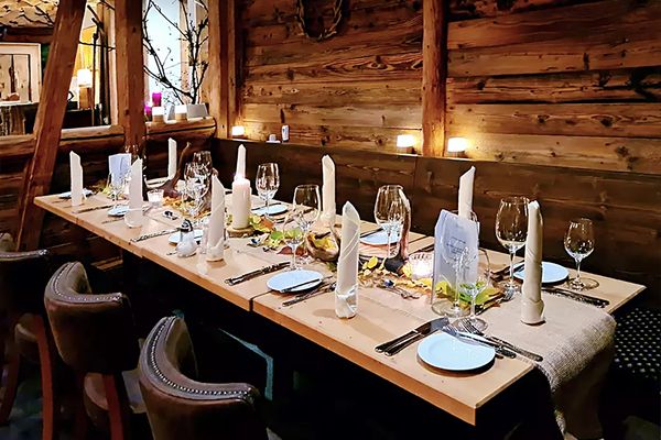 Blick in die rustikale Gasttube der Taufsteinhütte. Die Wände und Decken sind aus schwerem, dunklen Holz. Eine langgezogene Tafel, fein mit Weingläsern und Stoffservietten eingedeckt, mit schweren lederbezogenen Stühlen und einer Eckbank aus Holz ist das zenttrale Bildmotiv. Links dahinter kann man durch das offene Fachwerk in den angrenzenden Raum blicken.