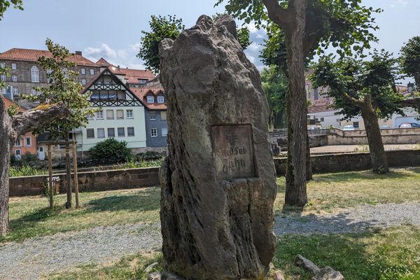 Das Bild zeigt einen Gedenkstein im Vordergrund. Dieser steht in der Jahnsallee in Kronach. Dahinter sieht man Bäume, sowie einen Teil der Altstadt.