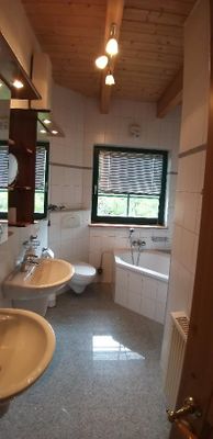 Badezimmer mit Eckbadewanne, WC, zwei Waschbecken, Spiegel und Holzdecke. Am Fenster befindet sich ein Innenrollo.
