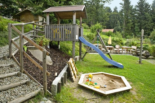 Garten-Spielplatz mit Sandkasten, Spielhaus mit Rutschbahn, Schaukel- und zusätzlichem Klettergerüst. Dahinter zwei Sitzgruppen aus Holz. Vor dem Spielplatz, der umgeben ist von hohen Bäumen, befindet sich ein kiesbestreuter Aufgang mit einem Holzzaun aus Rundlingen.