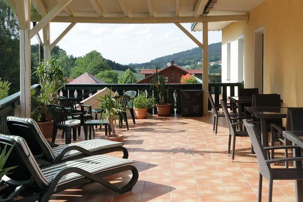Auf der überdachten Terrasse stehen viereckige Tische, Rattanstühle sowie Liegen und Gartenstühle mit grün gestreiften Auflagen und einige Grünpflanzen. Im Hintergrund der Blick auf Gebäude und bewaldete Hügel.