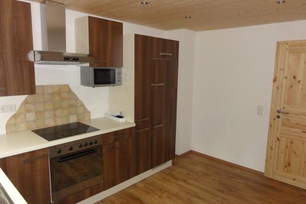 Küchenzeile mit heller Arbeitsplatte, dunklen Holztüren und einer Mikrowelle. In die helle Holzdecke sind mehrere Strahler eingebaut.