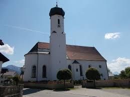 Der Betrachter blickt von der gegenüberliegenden Seite der Dorfstraße auf die weiße Dorfkirche mit der Friedhofsmauer.