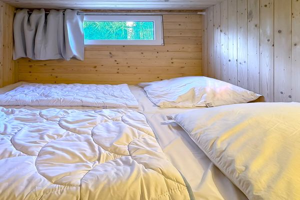 Schlafbereich mit zwei Matratzen und Kissen in einem vollständig mit Holz verkleideten Raum. Über den Matratzen befindet sich ein schmales Fenster mit Vorhang.