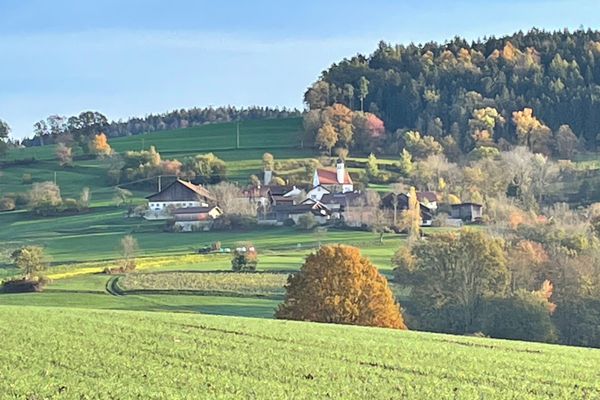 In einer hügeligen und herbstlichen Wald- und Wiesenlandschaft stehen mehrere Gebäude rund um eine kleine Kirche.