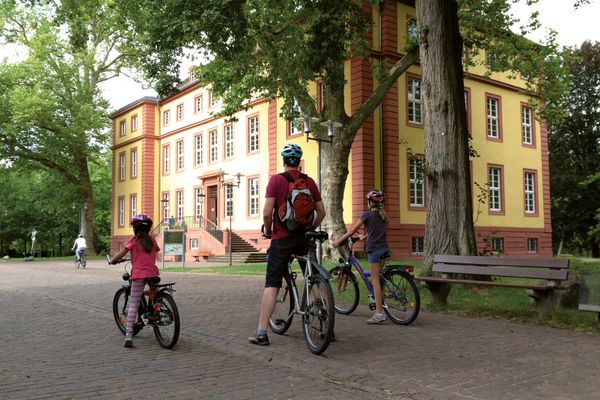 Drei Radfahrer, ein Mann und zwei Kinder, stehen vor dem mit Bäumen umwachsenen, klassizistischen Schlossbau. Das Gebäude ist groß und gelb mit Sandsteinkanten und Umrandungen der Fenster.